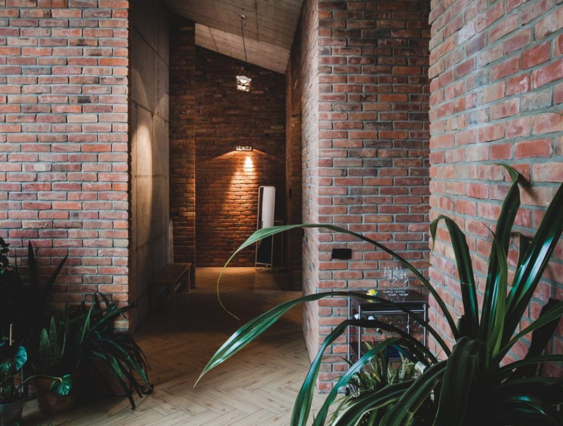 A hallway with rustic brick walls and wooden flooring, curated by renowned brick suppliers, features lush green plants like ferns and yucca. Ambient lighting creates a warm atmosphere, while an archway in the distance adds depth to the space.
