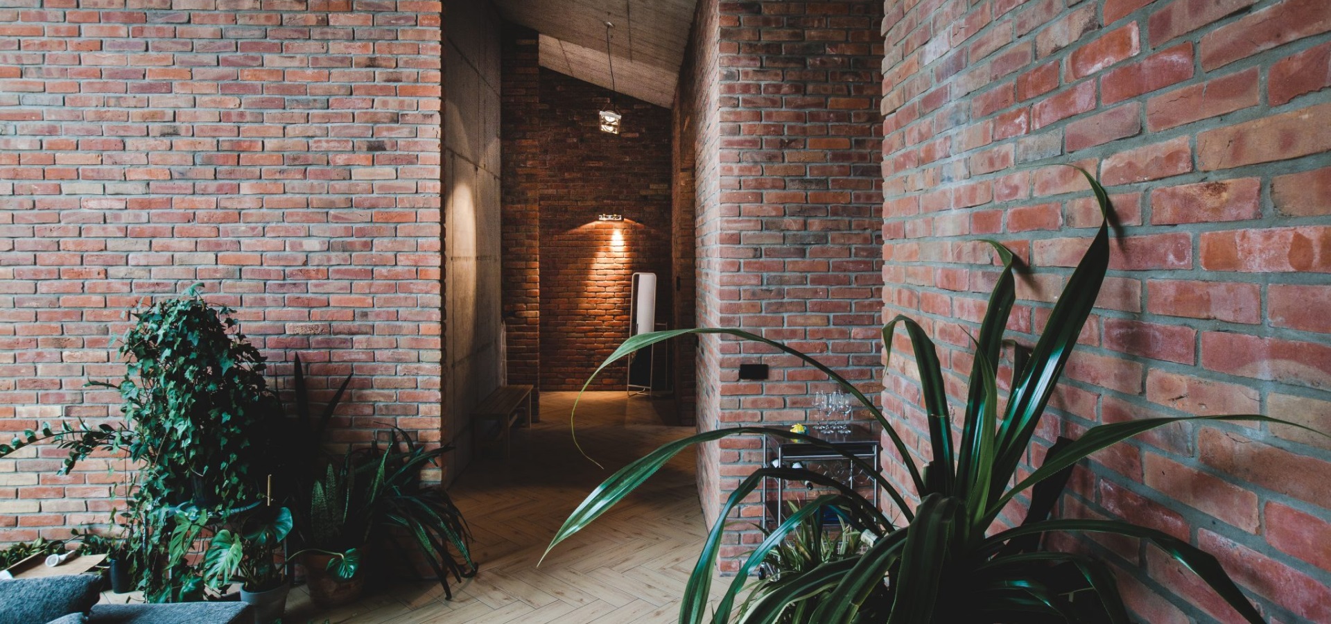 A hallway with rustic brick walls and wooden flooring, curated by renowned brick suppliers, features lush green plants like ferns and yucca. Ambient lighting creates a warm atmosphere, while an archway in the distance adds depth to the space.