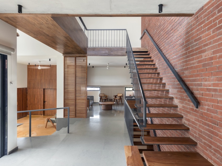 Modern interior featuring a wooden staircase with metal railing, seamlessly integrated with the brick-matching exposed wall on the right. Light-colored flooring complements the wooden panels and furniture in this spacious, open-plan area, where natural light floods through generous windows.