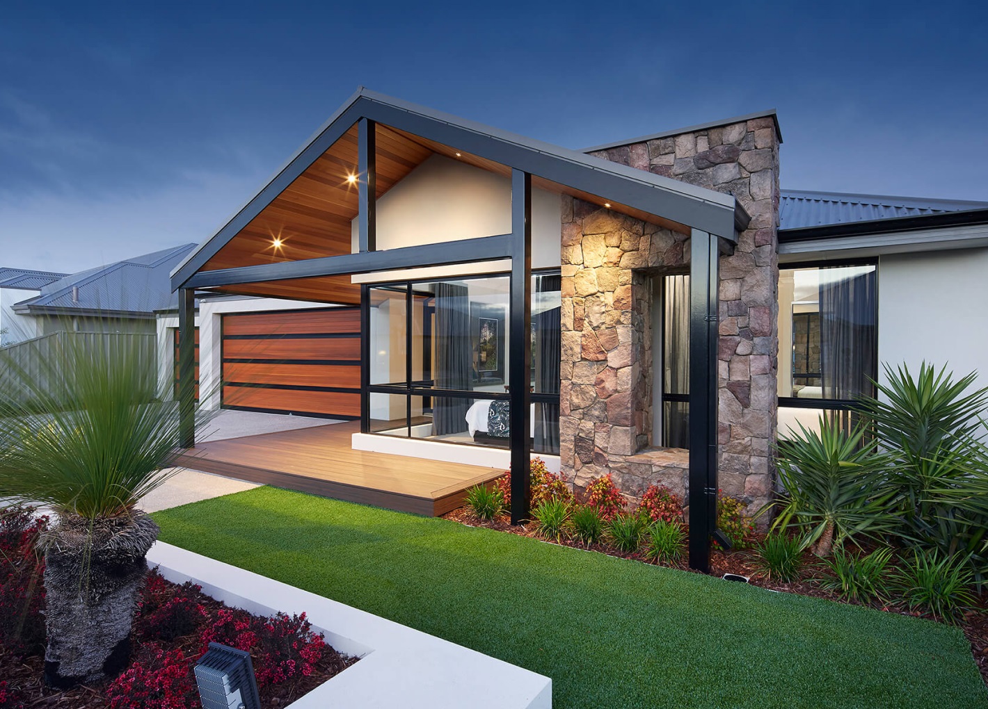 Modern one-story house with a mix of stone and wood facade, large windows, and a sloping roof. Enhanced by Architectural Designer Products, it features a well-lit entrance, manicured lawn, and neatly arranged plants, creating a warm and welcoming appearance at twilight.