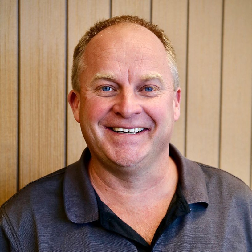 Brad Cairns, a smiling man with short light hair, stands confidently in front of a wooden panel wall, donning a dark polo shirt.
