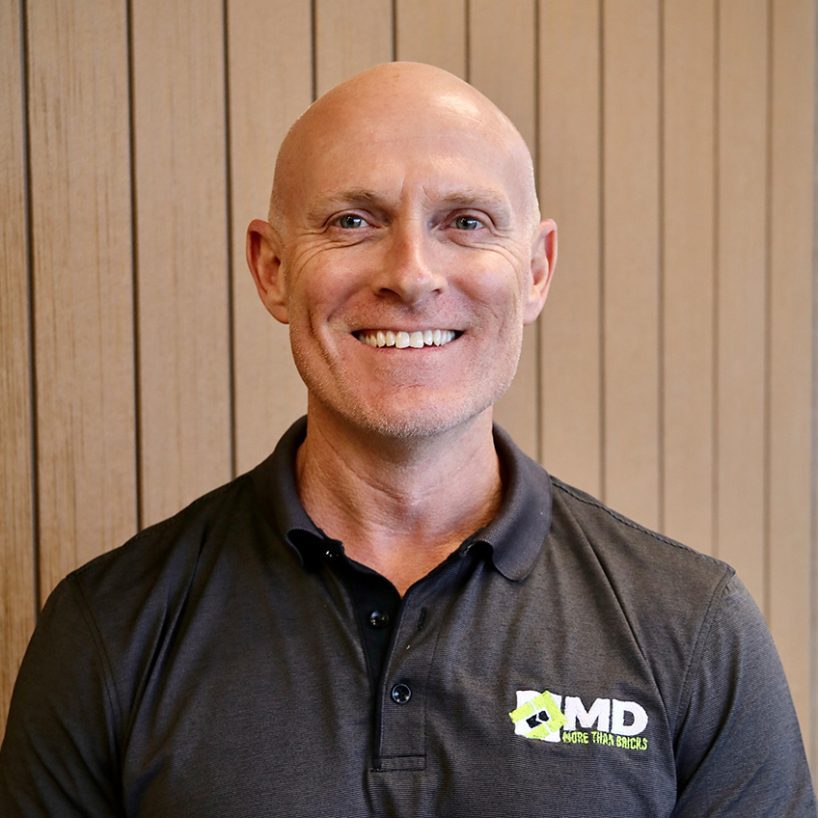 A smiling man with a bald head, identified as Jarrod Huxtable, wearing a dark polo shirt with an MD Brick Logo stands in front of a wooden panel wall.