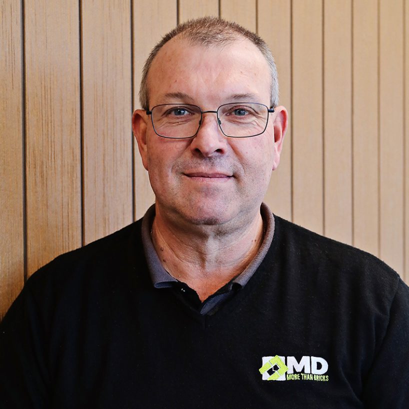 John Kirovski, a man with glasses and short hair, is standing against a wooden panel wall. He wears a black sweater proudly displaying the company logo that reads MD More Than Bricks.