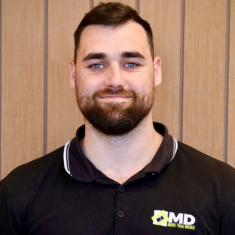 Mark Blundell, a man with short brown hair and a beard, stands confidently against a wooden panel background. He's wearing a black polo shirt with the logo 'MD MORE THAN BRICKS,' smiling warmly at the camera.