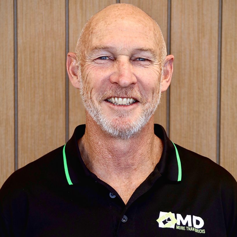 A smiling man, identified as Martin Driene, with a bald head and gray beard stands in front of a wooden panel wall. He sports a black polo shirt with green trim and a logo that reads MD More Than Bricks.