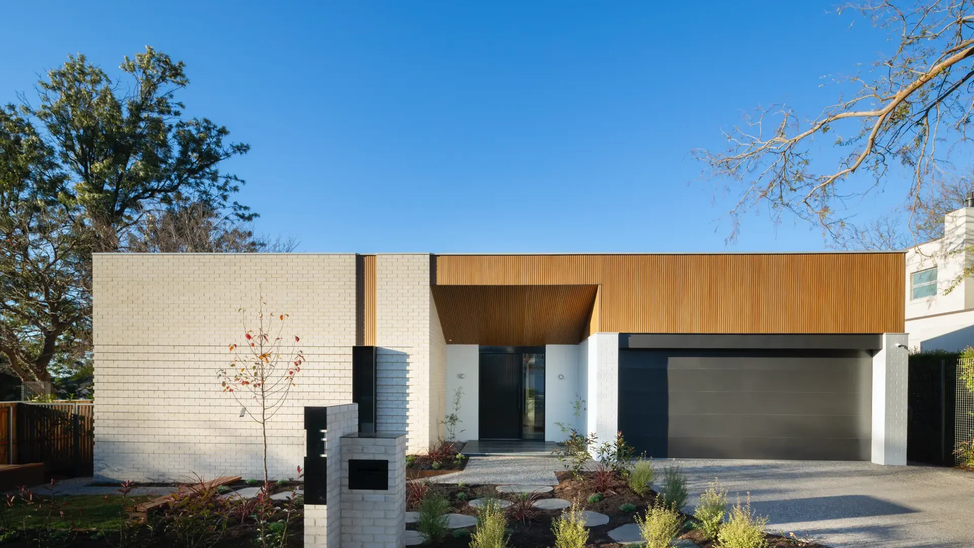 A modern, single-story house with a minimalist design, featuring a white brick exterior and wooden accents. The house has a black garage door, a small front yard with newly planted shrubs, and a clear blue sky in the background.