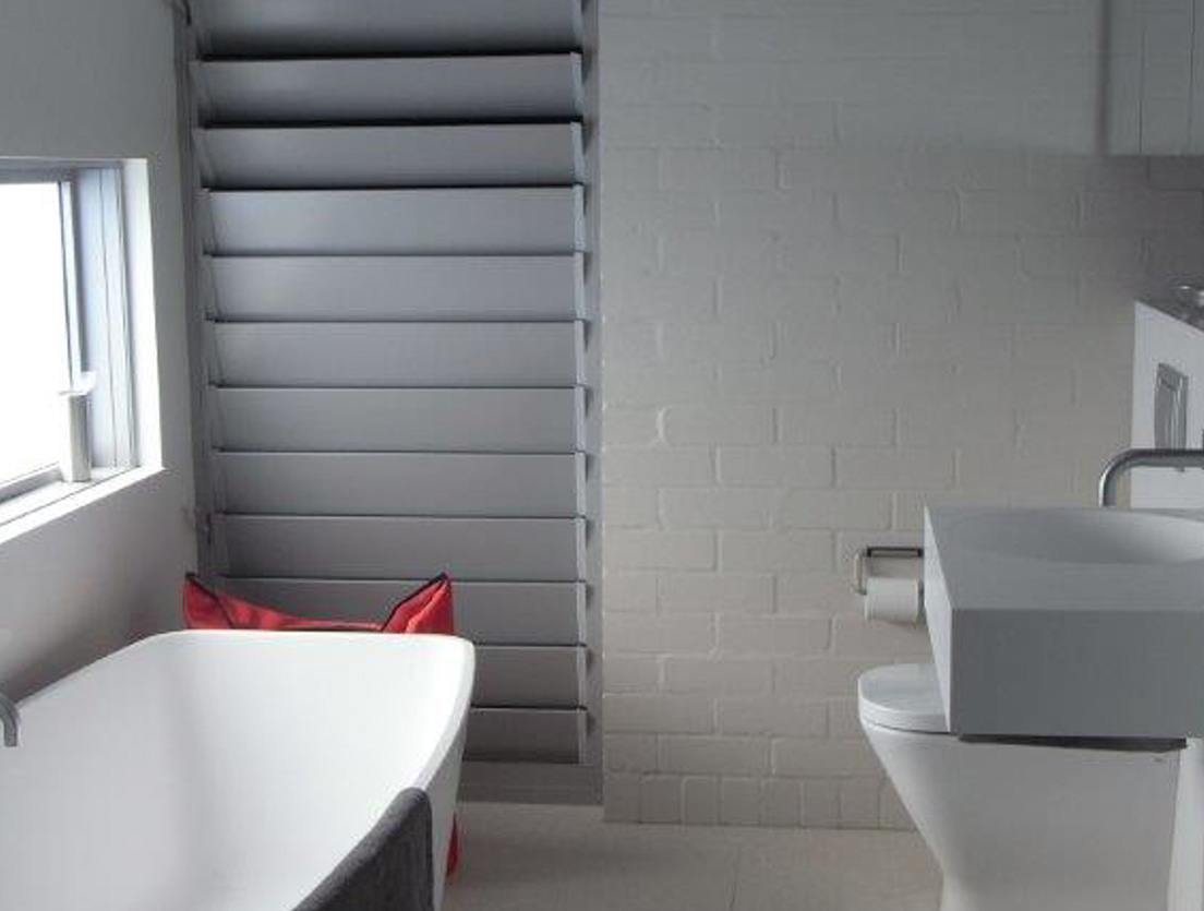 A modern bathroom features a white freestanding bathtub, a sleek gray sink, and a white toilet. The brick wall, painted with sustainable building materials, is white. A narrow horizontal window with blinds invites natural light in, while a red basket sits near the tub.