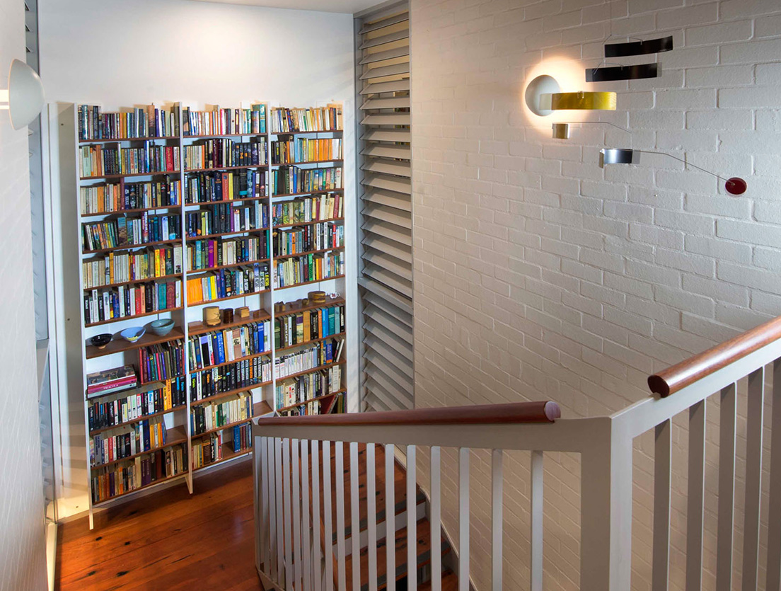 A staircase of sustainable wooden steps and a white railing leads to a landing with a tall bookcase filled with various books. The walls, made from sustainable white brick, feature a modern wall light fixture and a minimalist mobile art piece on display.