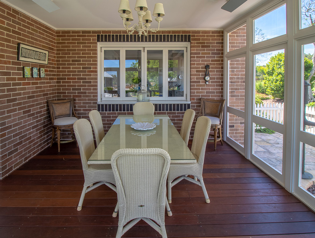 A sunlit room showcases a rectangular glass dining table surrounded by wicker chairs on a wooden floor crafted from sustainable building materials. Brick walls with windows and framed photos, a chandelier overhead, and an outdoor view complete the cozy setting.