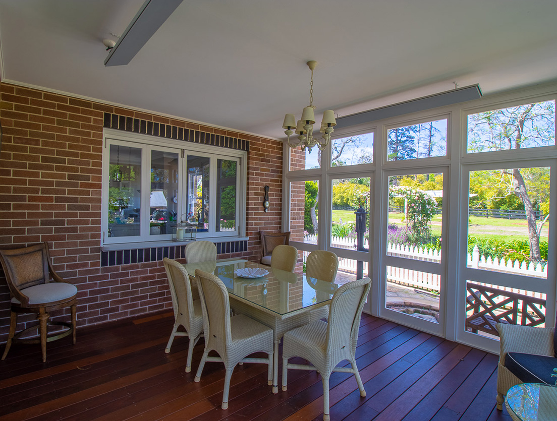 A sunlit dining room features a glass-top table with wicker chairs on a wooden floor crafted from sustainable building materials. Large windows and a door frame a view of a garden with trees and a white picket fence. A brick accent wall and chandelier add charm to the space.
