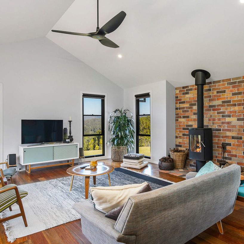 A modern living room showcases Architectural Designer Products with a sleek gray sofa, wooden coffee table, and TV on a white stand. A black ceiling fan graces the high ceiling above. A brick wall features a fireplace and a plant near expansive windows revealing a lush landscape.