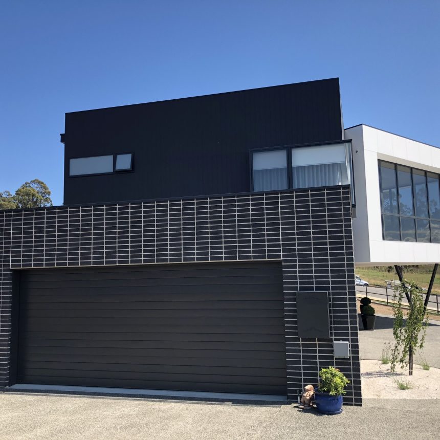 A contemporary two-story house showcases a stylish black and white exterior, featuring Designer Smooth Graphite tiles on the garage and ground level. The second floor highlights expansive windows and a cantilevered design, while a blue potted plant adorns the front under the clear blue sky.