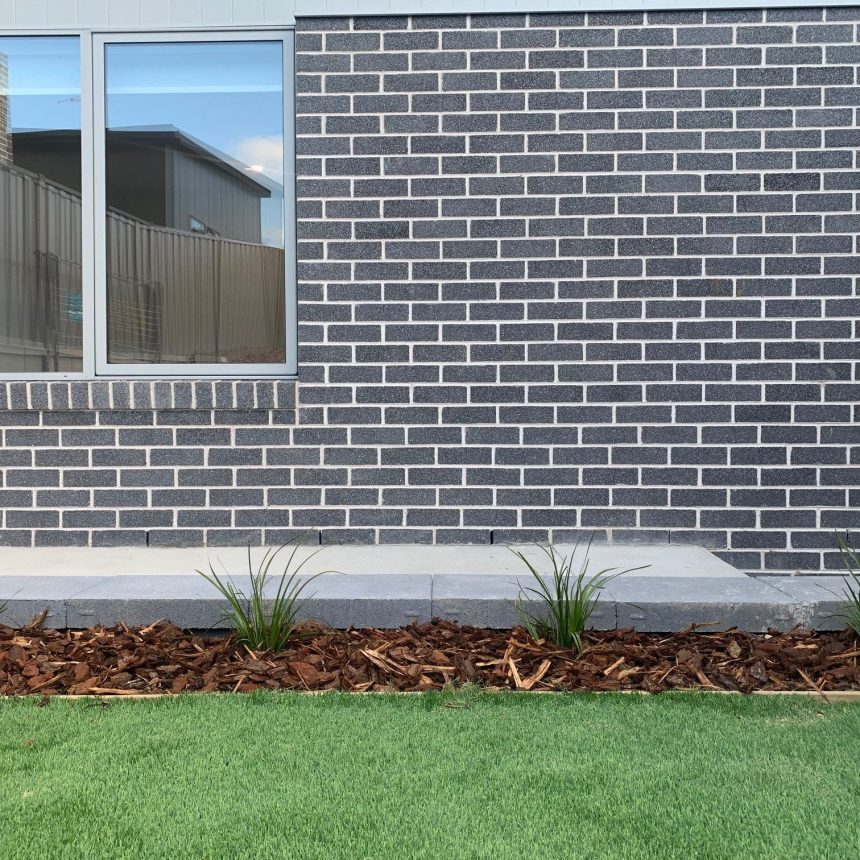 A modern brick wall with a large window sits above a small garden bed, its mulch arranged artfully and punctuated by refined plants. In front, a strip of enhances the scene’s bright and outdoor tones.