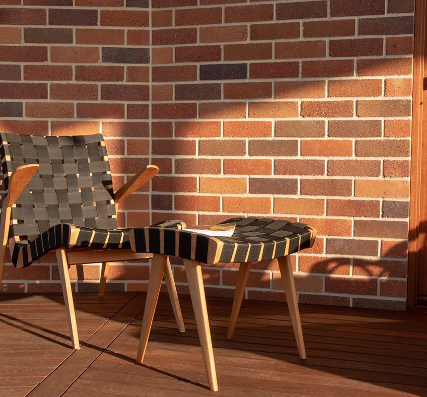 A Brick wall crafted with the Lincoln Classic Mottle Blend pattern, accompanied by a matching ottoman and chair, sits on a wooden deck against a brick wall. Bathed in warm sunlight, the seat holds an open book on the ottoman, while a clear glass door mirrors the outdoor scenery.