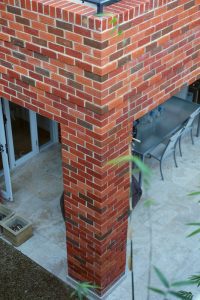 The Textures Red No Aris brick corner of a building highlights a glass door that opens to an inviting patio area. On the tiled floor, a table with chairs is encircled by plants, while the vibrant red accents bring warmth and style to create an attractive outdoor seating space.
