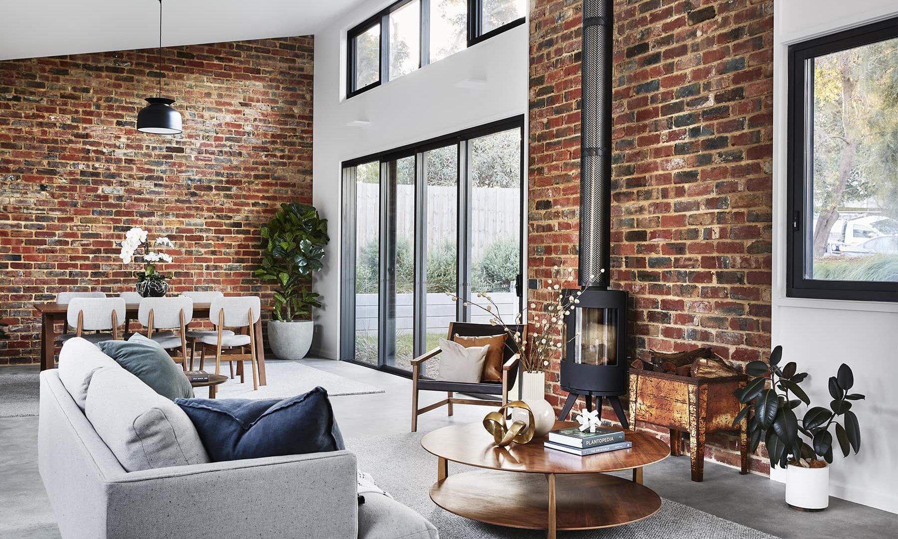 Inside a house with an exposed brick wall with glass doors in the centre of the wall and a black fireplace with a chimey running up the exposed brick wall and a light coloured lounge and a small wooen coffee table