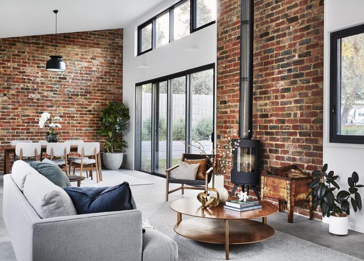 Inside a house with an exposed brick wall with glass doors in the centre of the wall and a black fireplace with a chimey running up the exposed brick wall and a light coloured lounge and a small wooen coffee table