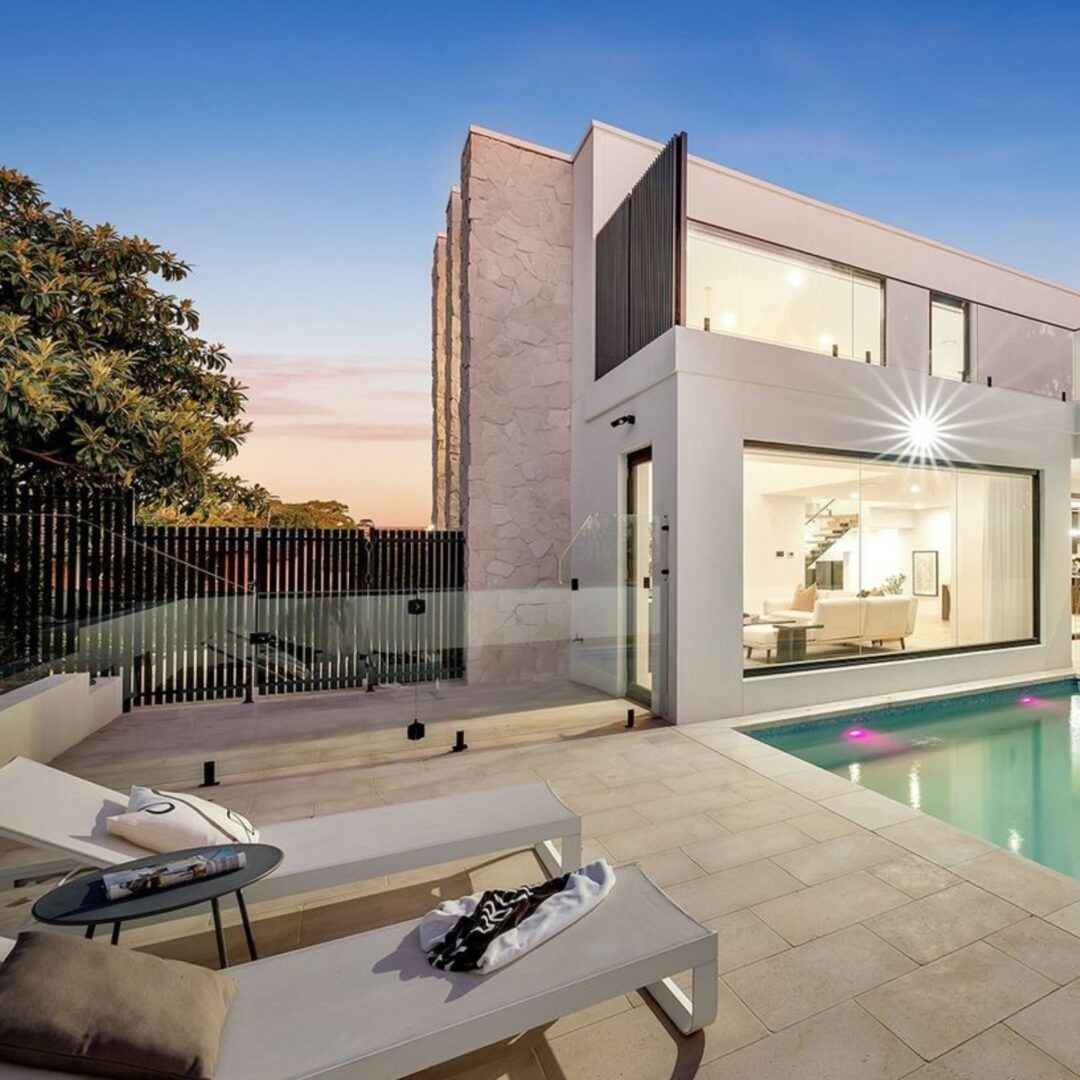 This modern two-story house features a glass-walled pool surrounded by Sinai Pearl S/blasted & Tumbled Paver. Two lounge chairs on the tiled patio complement stone accents and large windows that reflect the sunsets warm glow.
