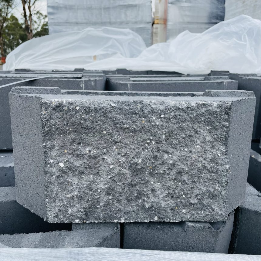 Displayed outdoors is a stack of Norfolk Basalt, showcasing its distinct rugged charm with gray concrete blocks having a rough texture. Several blocks are protected by clear plastic sheeting, and trees can be seen in the background.