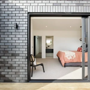 At La Palomo Azul, a modern bedroom with a pink bedspread is seen through large sliding glass doors in a black brick wall, while a black chair stands outside on the wooden deck, adding to the bright, minimalistic style.