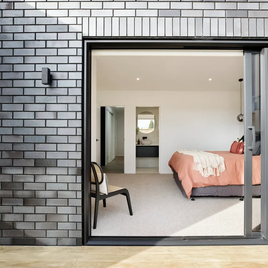 At La Palomo Azul, a modern bedroom with a pink bedspread is seen through large sliding glass doors in a black brick wall, while a black chair stands outside on the wooden deck, adding to the bright, minimalistic style.