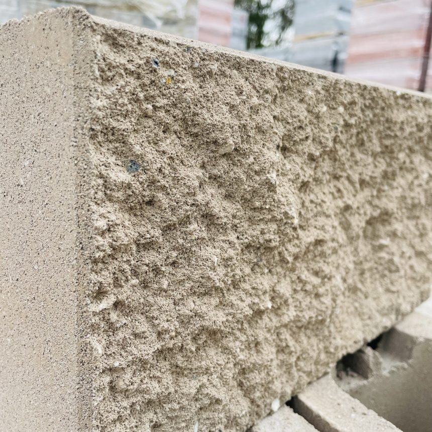 Close-up of a Split Face Bushrock concrete block showcasing its rough, uneven surface. Several similar blocks are stacked in the background, reminiscent of bushrock, with a hint of greenery visible, indicating an outdoor construction site setting.