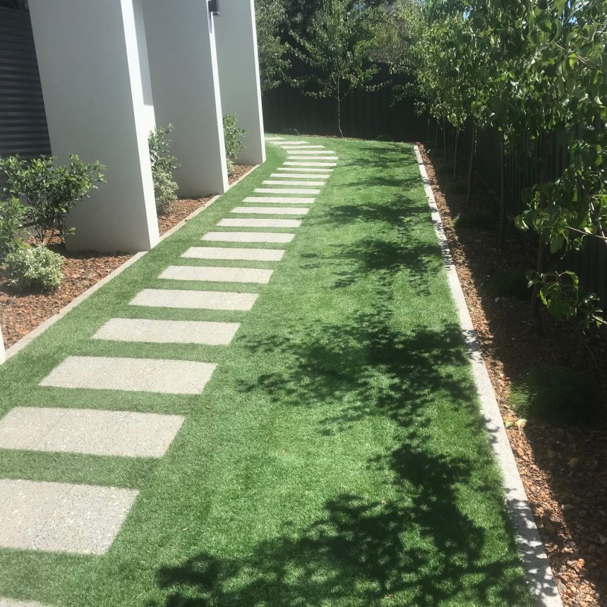 A narrow garden boasts a neat pathway of Refined Ebony Pebble tiles set in artificial grass. The path elegantly curves beside a building, while small trees and plants line the mulched border on the right, casting their shadows on the grass.