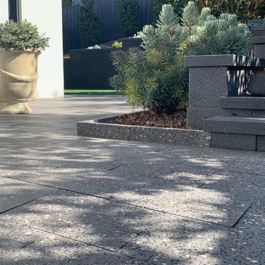 A contemporary outdoor patio adorned with Classic Stone's Refined Ebony Pebble tiles, showcasing a spacious decorative planter and a raised garden bed filled with vibrant green shrubs. Stone steps, crafted from the same elegant material, lead to another section that is partially visible in the background.