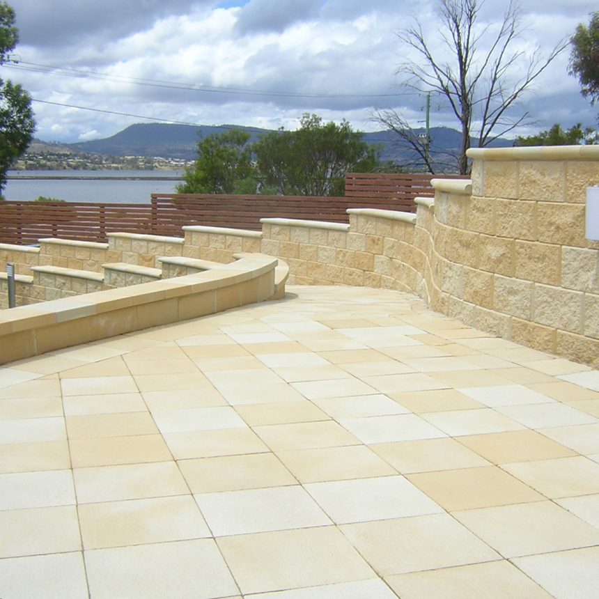 A spacious outdoor patio features Classic Stone Smooth Limestone tiles and curving retaining walls. It offers a view of a body of water encircled by trees and hills beneath a cloudy sky.