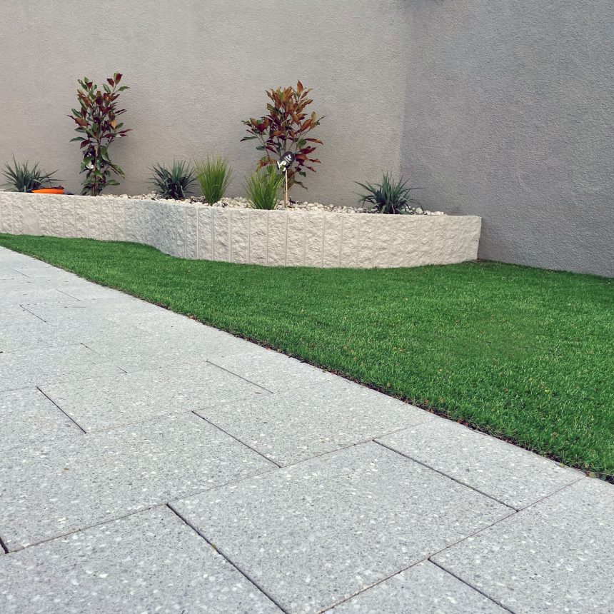 A neatly landscaped yard featuring a rectangular patch of green grass next to a light gray paved pathway. In the corner, there's a stone planter filled with small plants and Classic Stone Refined Mist Pebble against a gray wall.