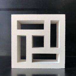 The Breeze Block Hindu White features a geometric design resembling a solid grey rectangular grid with variously sized and positioned cutouts, creating an abstract sculpture. Its stark white color contrasts beautifully against the dark background.