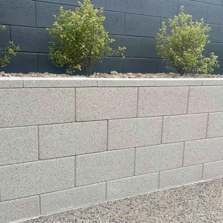 A wall constructed from DIY Coloured Blocks Mist ECO is adorned with a line of small green bushes on top. It stands against a darker backdrop, with a gravel bed nestled between the blocks and the bushes. In the foreground, there is a surface with a pebble texture.