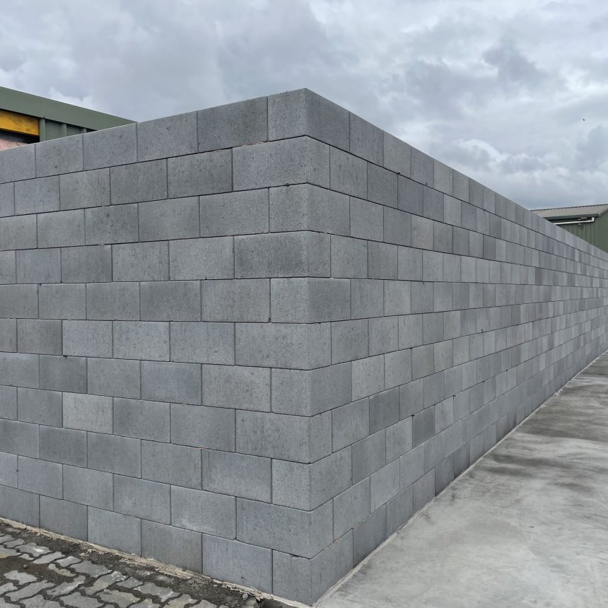 A long L-shaped wall of DIY Coloured Blocks Mist ECO extends along a paved area beside a green metal building under a misty sky.