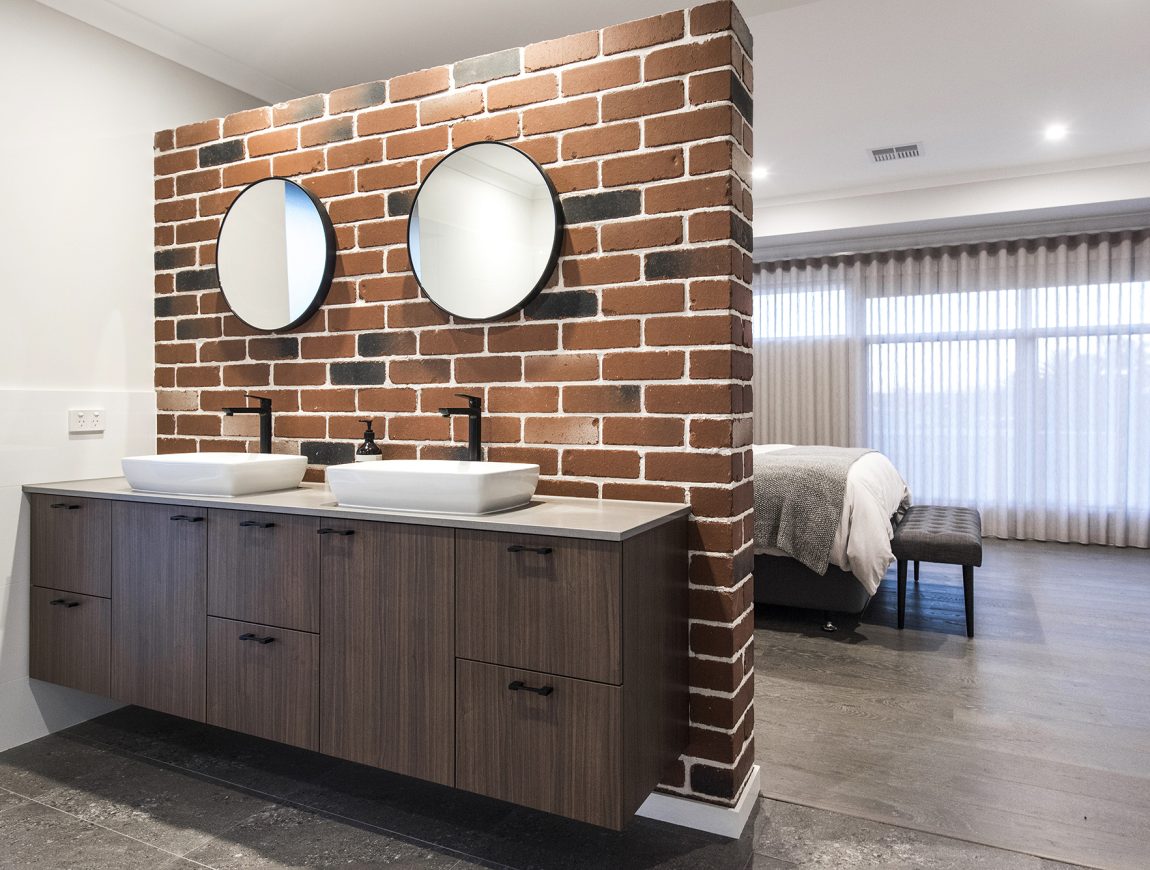 Modern bathroom with a double vanity featuring two round mirrors and vessel sinks on a wooden cabinet. The room boasts a Midland Bricks accent wall and opens to a bedroom with a bed and sheer curtains. The flooring is dark, and the space is well-lit.
