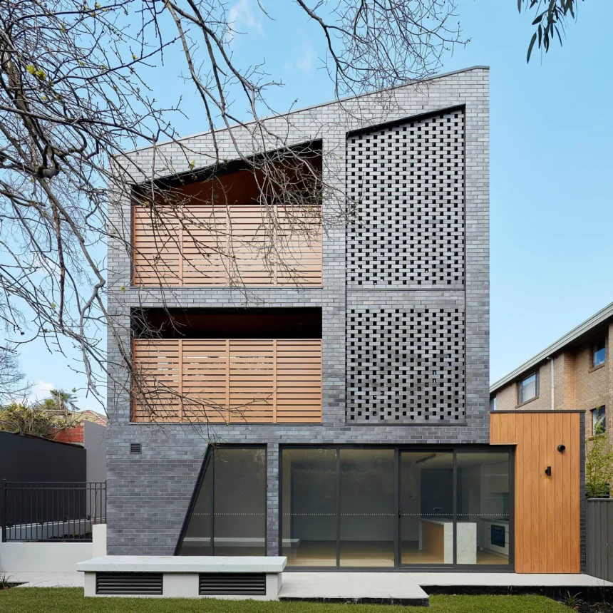 La Palomo Azul is a modern three-story home featuring dark gray brick, large glass windows, wooden panels, and a patterned brick facade set behind a green lawn and neighboring buildings.