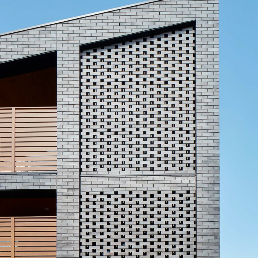La Palomo Azul features a modern facade with light gray brickwork and a lattice-style brick screen. Wooden slats on the left cover balcony openings, and the geometric design stands out against a clear blue sky.