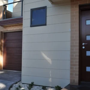 Modern house exterior clad in Stratum™ light beige panels, complemented by a dark brown door with four frosted glass panels, matching garage door, small upper window, and minimalist landscaping with white stones and small bushes.
