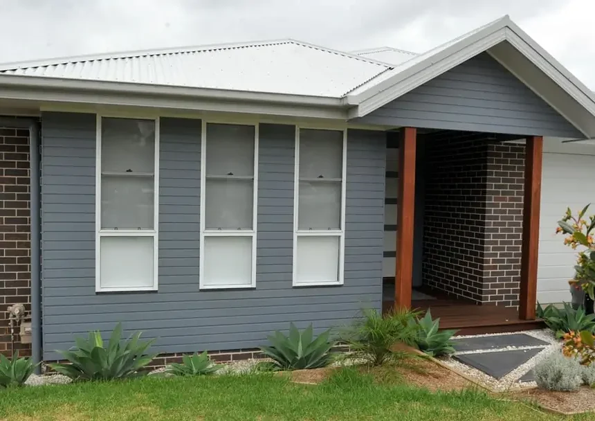 A modern single-story Stratum™ home with grey siding, white-framed windows, a white metal roof, and brown brick accents by the entrance. Thoughtful landscaping and small plants boost the front yards curb appeal.