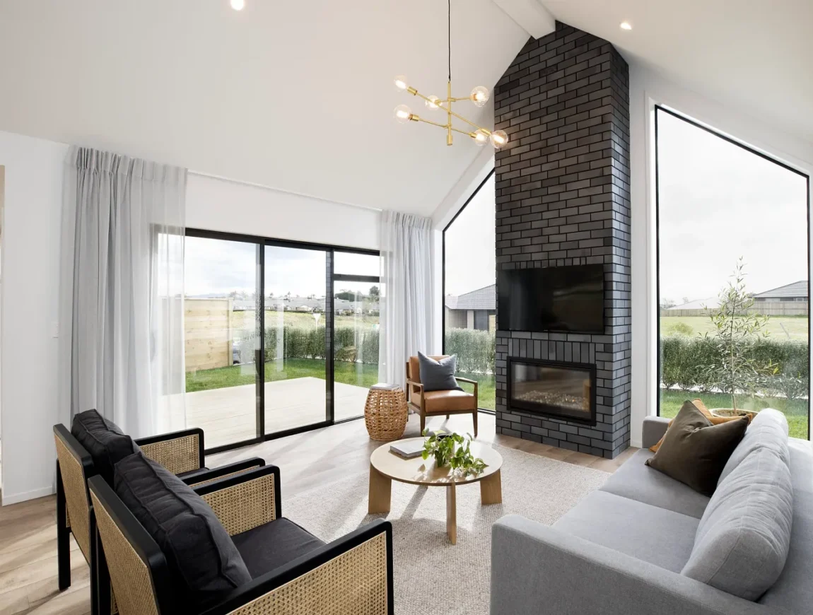 Modern living room with large windows, black brick fireplace, mounted TV, gray sofa, and two La Palomo Azul chairs with cane sides beside a round wooden coffee table on light wood floors and sheer curtains leading to the patio.