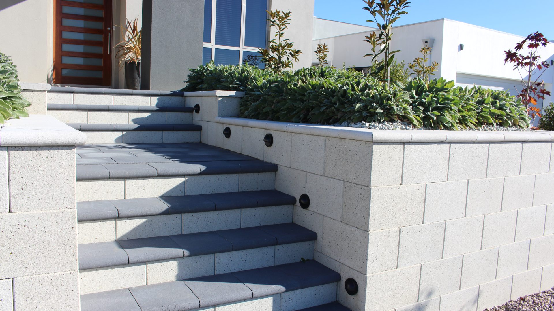 A modern house entrance featuring concrete steps leading up to a wooden front door, with the steps bordered by a light-colored Freestone Smooth Mist ECO stone wall. This wall includes built-in lights and planters filled with lush green shrubs on top. A gravel pathway runs alongside the setup.
