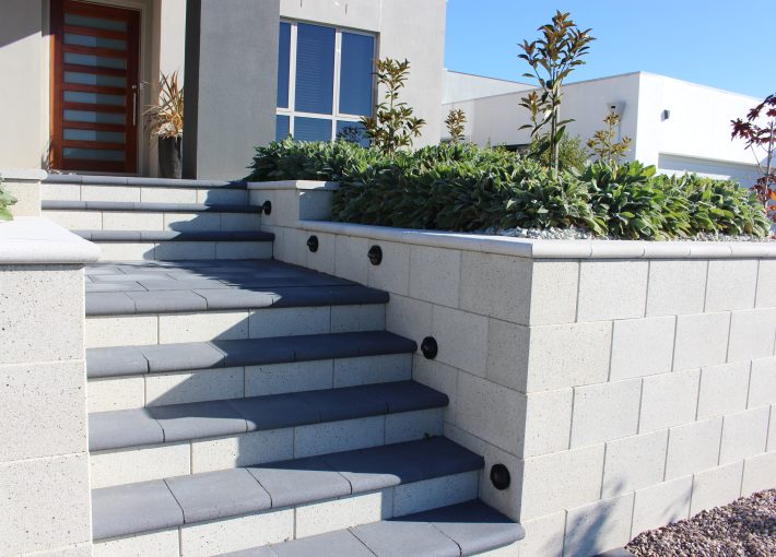 A modern house entrance featuring concrete steps leading up to a wooden front door, with the steps bordered by a light-colored Freestone Smooth Mist ECO stone wall. This wall includes built-in lights and planters filled with lush green shrubs on top. A gravel pathway runs alongside the setup.