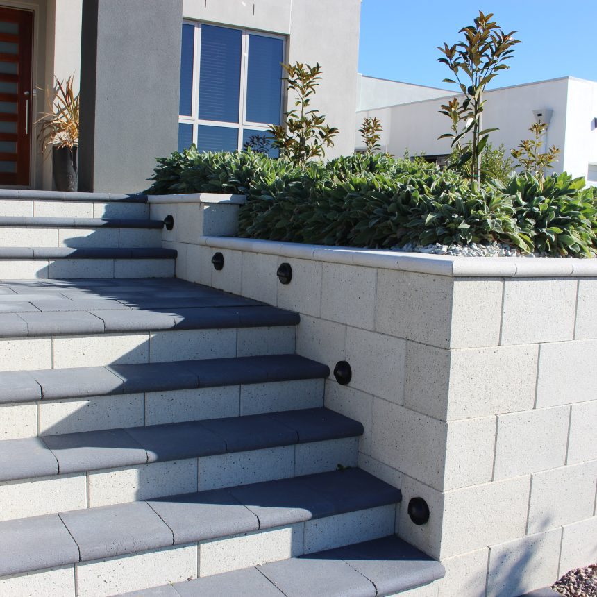 A modern house entrance featuring concrete steps leading up to a wooden front door, with the steps bordered by a light-colored Freestone Smooth Mist ECO stone wall. This wall includes built-in lights and planters filled with lush green shrubs on top. A gravel pathway runs alongside the setup.