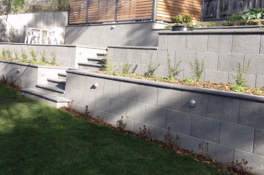 Terraced garden featuring Freestone Exposed Ebony ECO retaining walls and steps. Each tier is adorned with small plants and shrubs. At the top level, there is a wooden fence housing a white patio table and chairs, with a green lawn in the foreground.