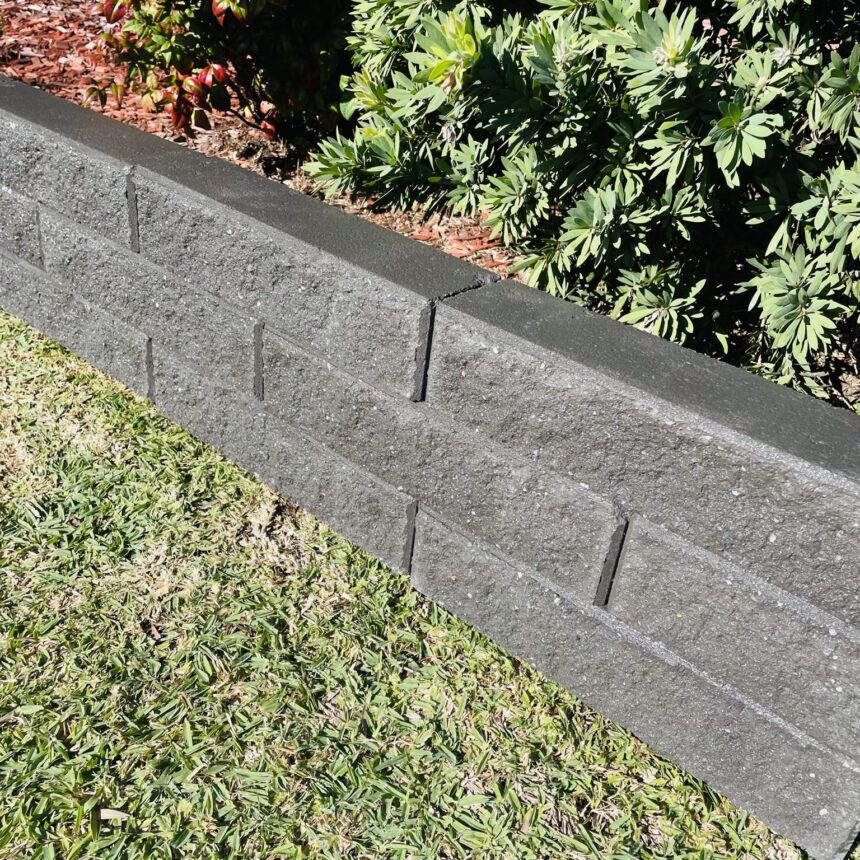 A low Broughton Garden Edge Basalt retaining wall in dark gray separates a grassy lawn from a garden bed with green shrubs and red-tipped leaves. Bright sunlight highlights the wall and casts shadows on the grass.