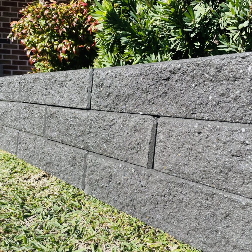 A close-up of a low, dark gray Broughton Garden Edge Basalt wall borders green grass with leafy shrubs behind it. In the background, there’s a brick building and part of a porch.
