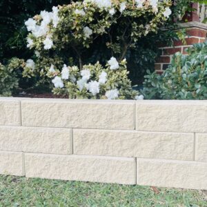 A short beige retaining wall made with Broughton Garden Edge Basalt stands before green grass, with a white-flowered bush and dense foliage in the background. A brick structure appears on the right.