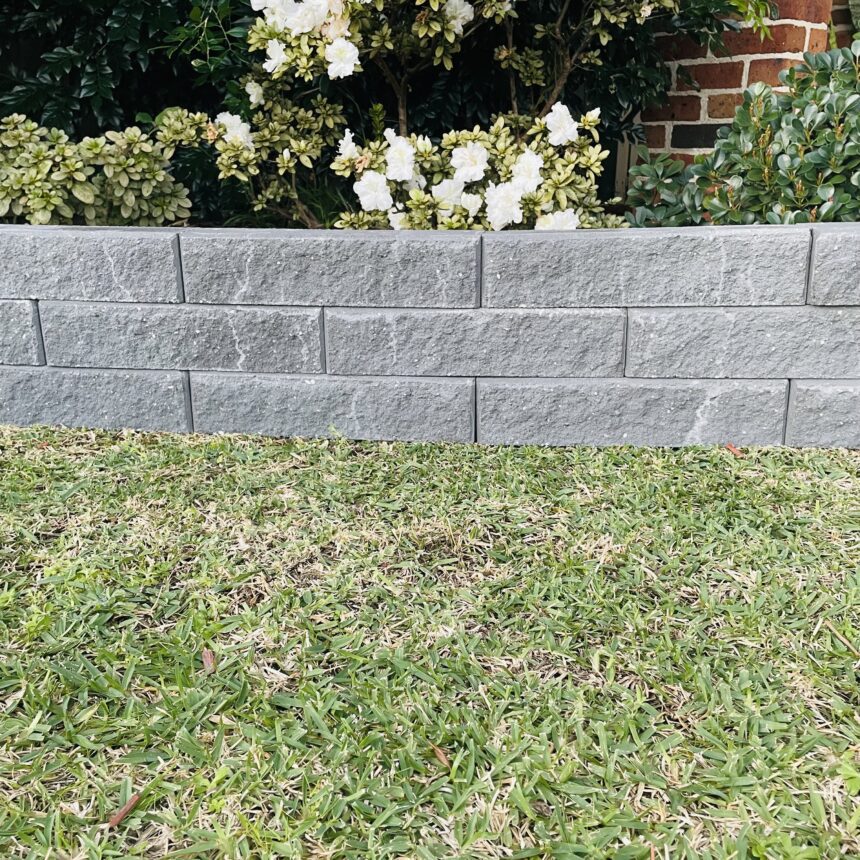 A low Broughton Garden Edge Basalt wall lines the lawn, with white flowering bushes behind it and a glimpse of red brick structure on the right.