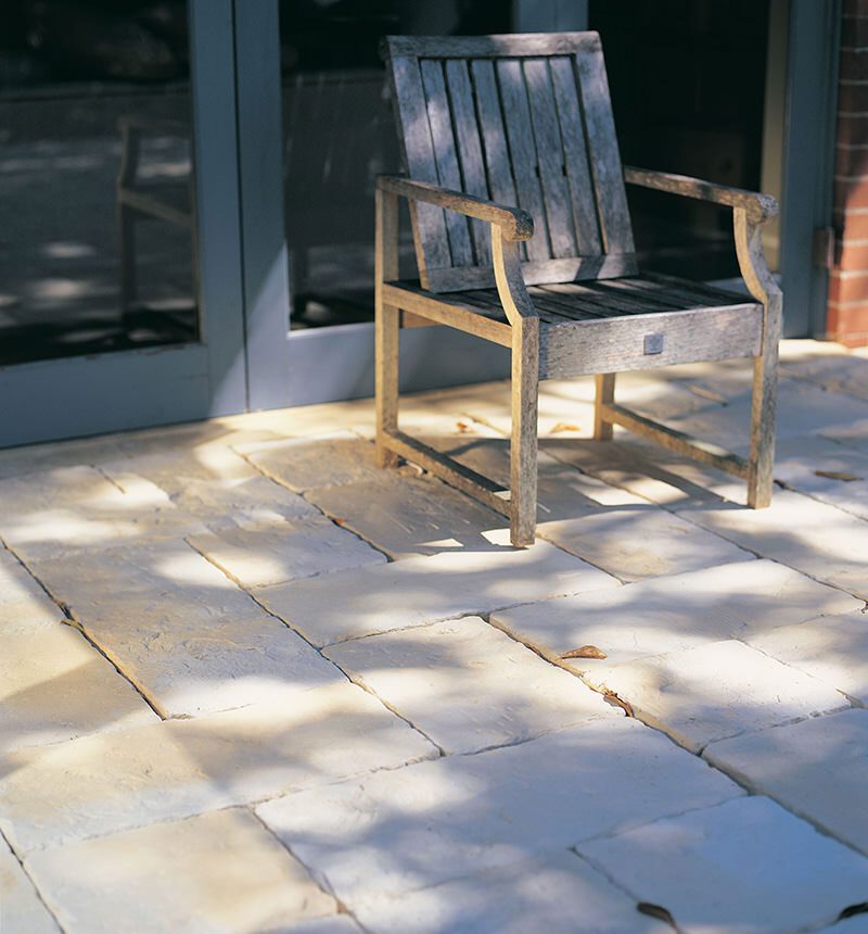 The Abode Iced Coffee pairs perfectly with a wooden chair on a patio of large, light stone tiles. Leaf shadows dance on the ground, crafting a serene retreat. A glass door and brick wall complete the tranquil setting.