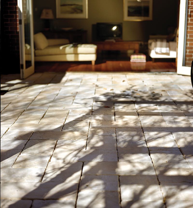 Sunlight plays on the stone patio, casting a trees shadow. Beyond, a cozy living room with a sofa, armchair, and wall art awaits. Open glass doors bridge indoors and outdoors, inviting one to enjoy an Abode Iced Coffee in the refreshing ambiance.