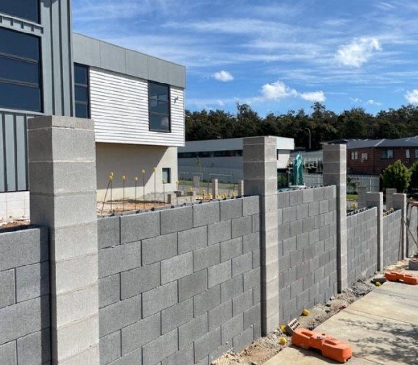 A partially constructed block wall, featuring DIY Coloured Blocks Ebony ECO for an eco-friendly touch, includes scattered colored blocks in shades of gray and other hues. It stands in front of a modern building with construction tools and materials strewn around. Two vertical pillars rise at the far end, as a worker in a green shirt diligently continues the project in the background.